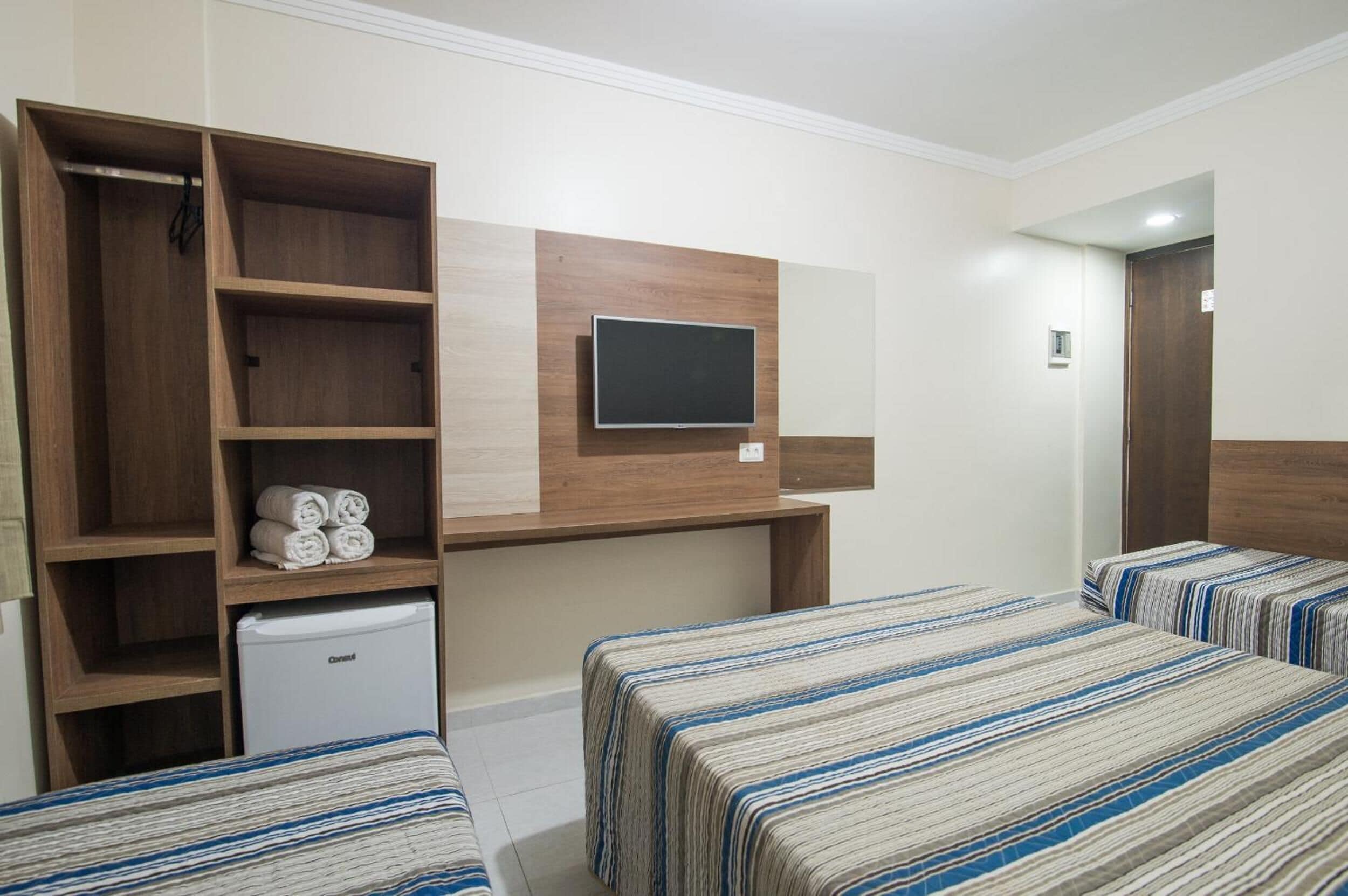 A hotel room with two beds, a TV mounted on the wall, a wardrobe, and a mini-fridge.