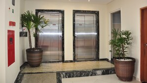A lobby with two elevators, a red emergency phone, and two large potted plants. - Hotel Guarumar (Guarujá)