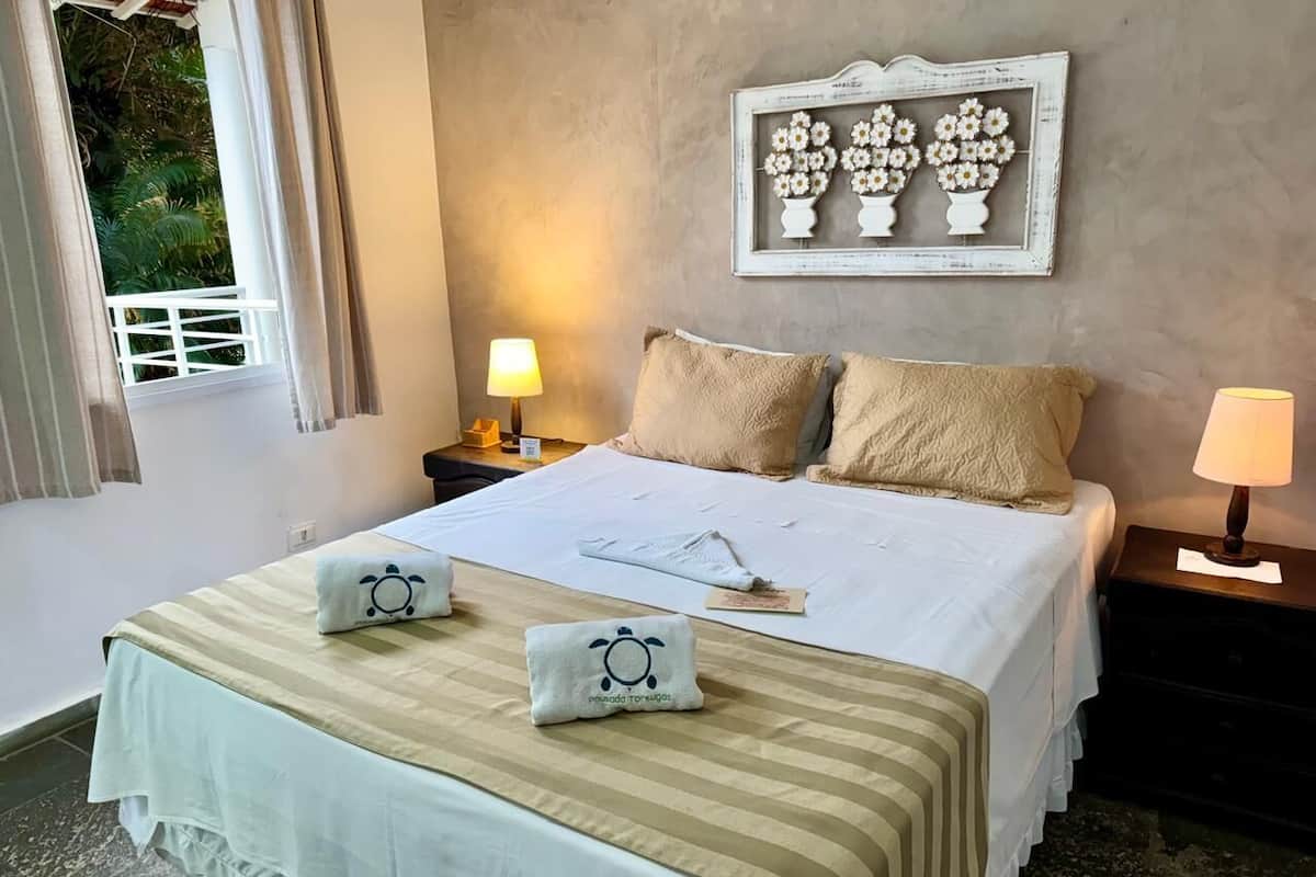 Cama bem arrumada com colcha listrada, dois travesseiros e dois travesseiros decorativos com desenho de tartaruga. Há um quadro com flores na parede acima da cama, e duas luminárias de cabeceira.