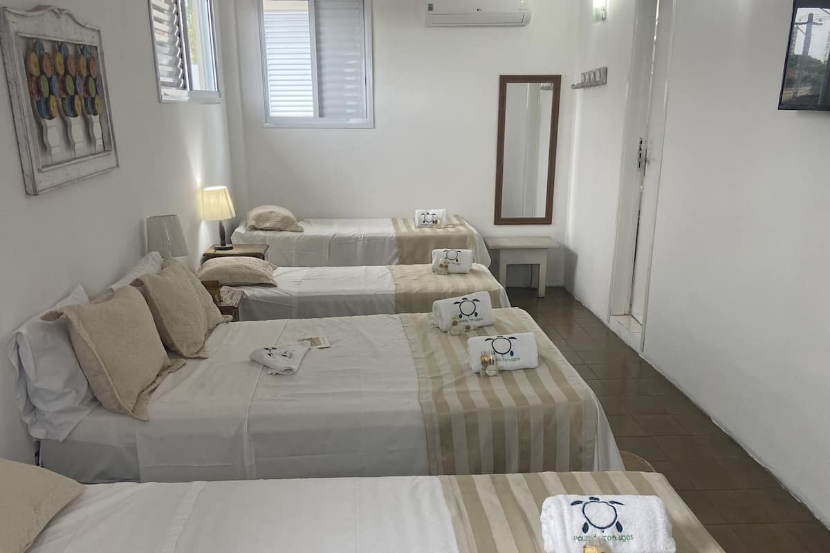 Quarto de hotel com três camas, cada uma com colcha listrada e toalhas com logotipo. Há uma janela com persianas, um ar condicionado e um espelho na parede.