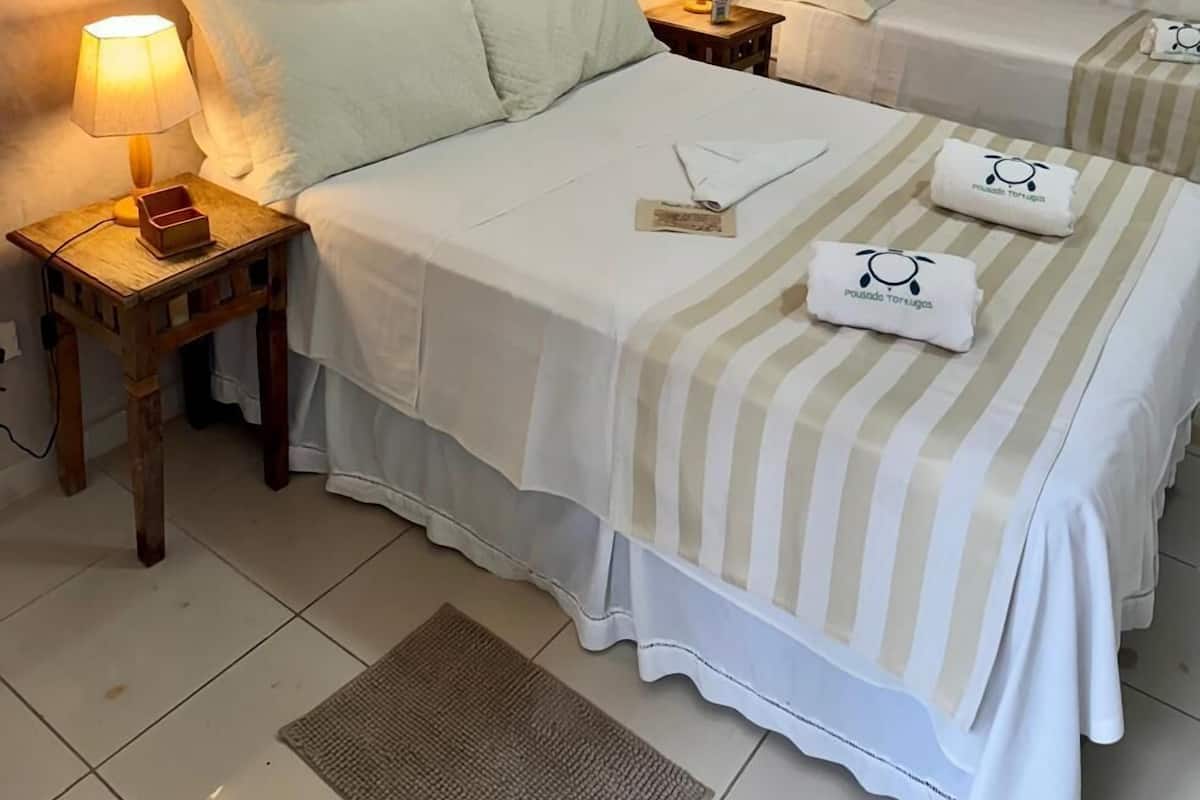 Quarto de hotel com duas camas, cada uma com colcha listrada e um par de toalhas com o logotipo. Há uma pintura de flores na parede, uma mesa de cabeceira com abajur e uma janela com cortinas.