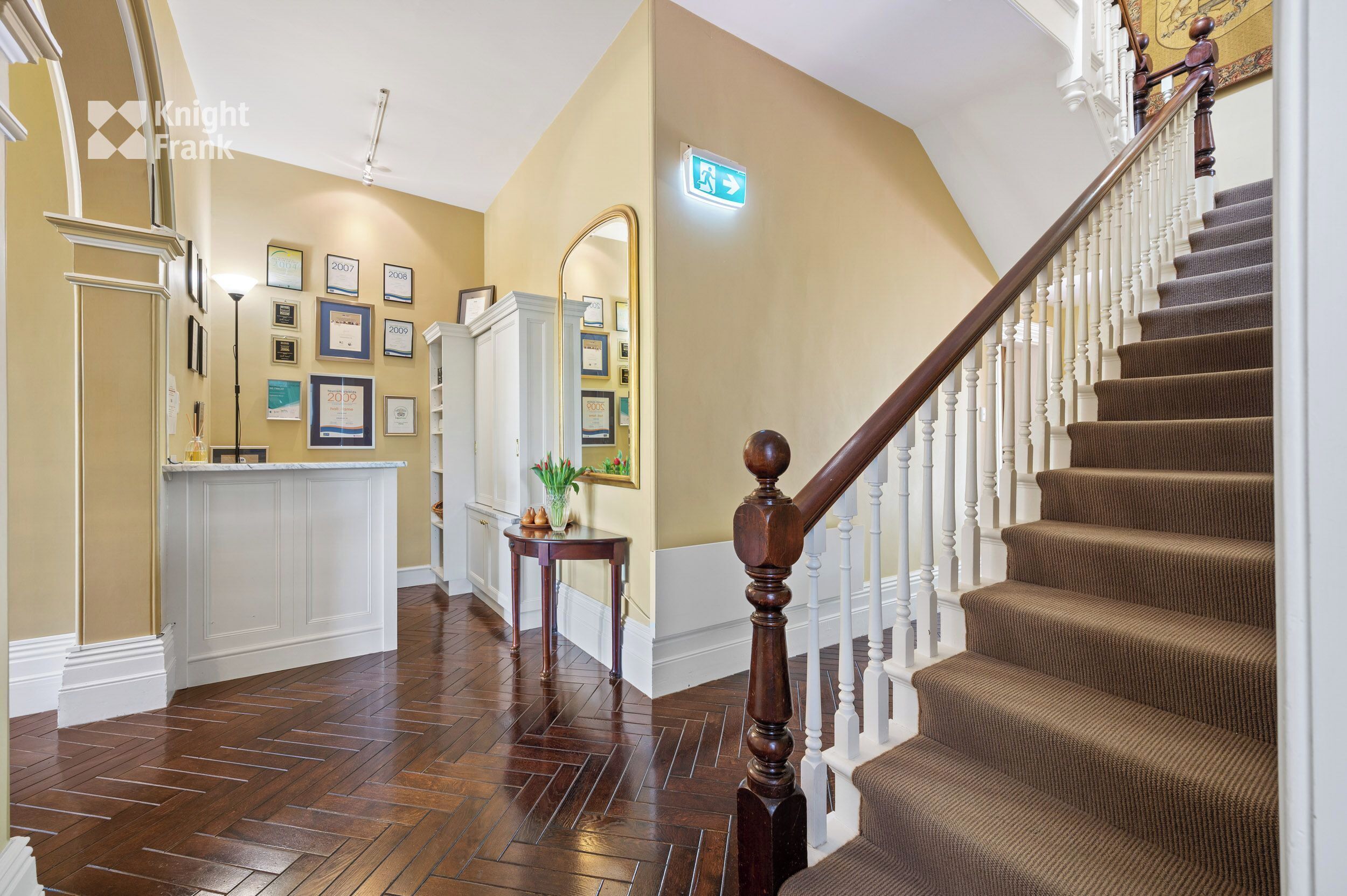 A hotel lobby with a wooden staircase, a reception desk, framed awards on the wall, and a mirror.