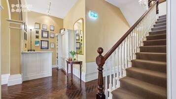 A hotel lobby with a wooden staircase, a reception desk, framed awards on the wall, and a mirror.