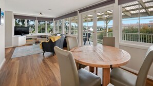 A spacious living area with a round wooden table and chairs, a sofa, and a TV. Large windows offer views of the surrounding area. - Clydesdale Manor (Sandy Bay)