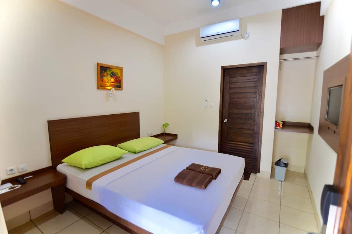 A hotel room with a bed, green pillows, a bedside table with a remote control, a small plant, and a painting on the wall.