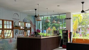 A modern reception area with a wooden reception desk, large windows offering a view of greenery, and a ceiling fan. - Central Inn (Senggigi)