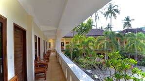 A hotel corridor with wooden benches and a view of a garden with palm trees and plants. - Central Inn (Senggigi)