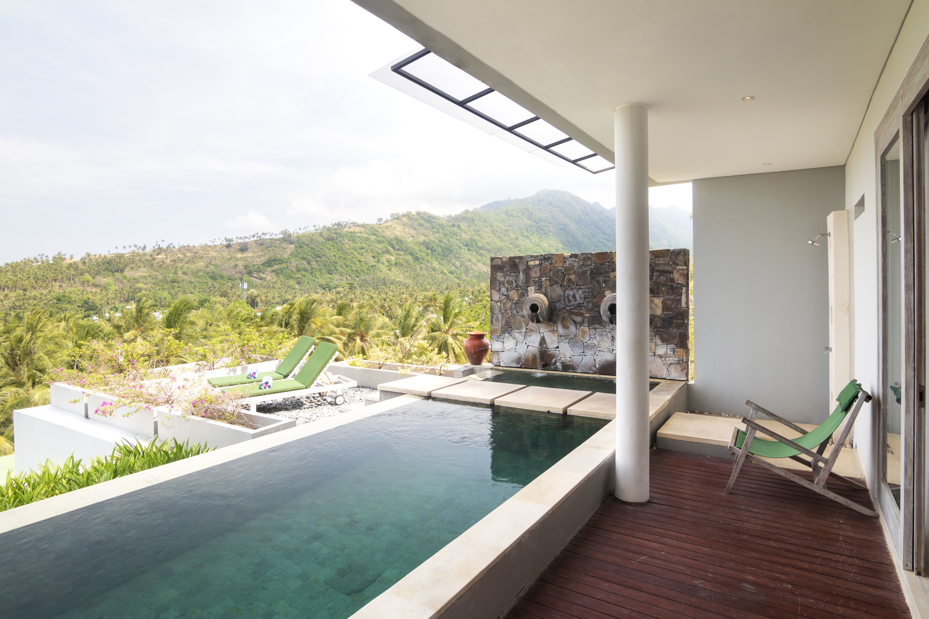 A modern outdoor pool area with a view of a lush hillside, a stone wall, and a wooden deck.
