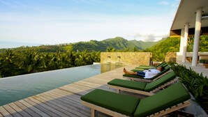 Une terrasse en bois avec des fauteuils de jardin confortables aux coussins verts, donnant sur une piscine, avec une chaîne de montagnes en arrière-plan.