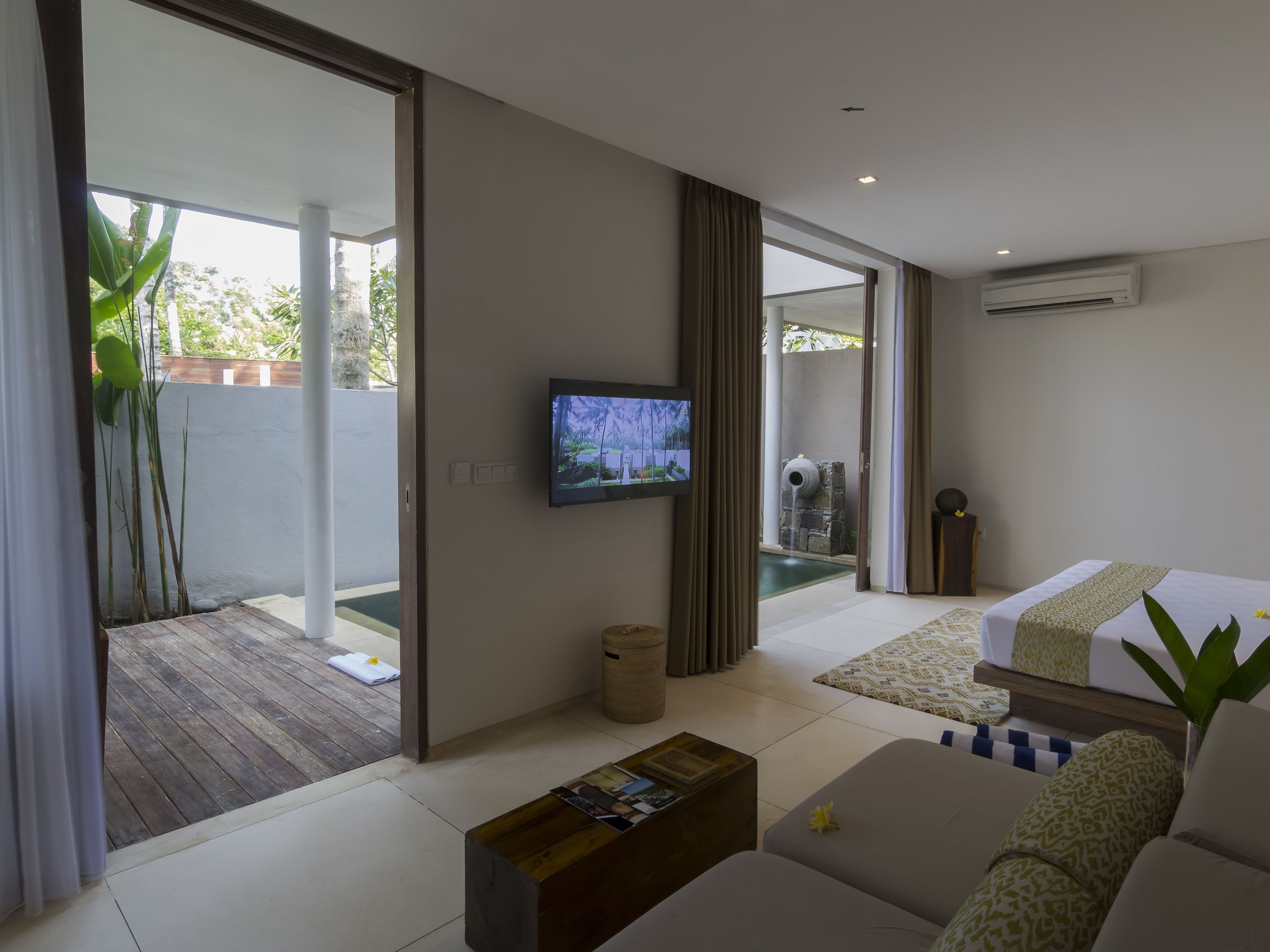 A modern hotel room with a large sliding glass door leading to a pool area, a flat-screen TV mounted on the wall, a wooden coffee table, and a bed with patterned bedding.