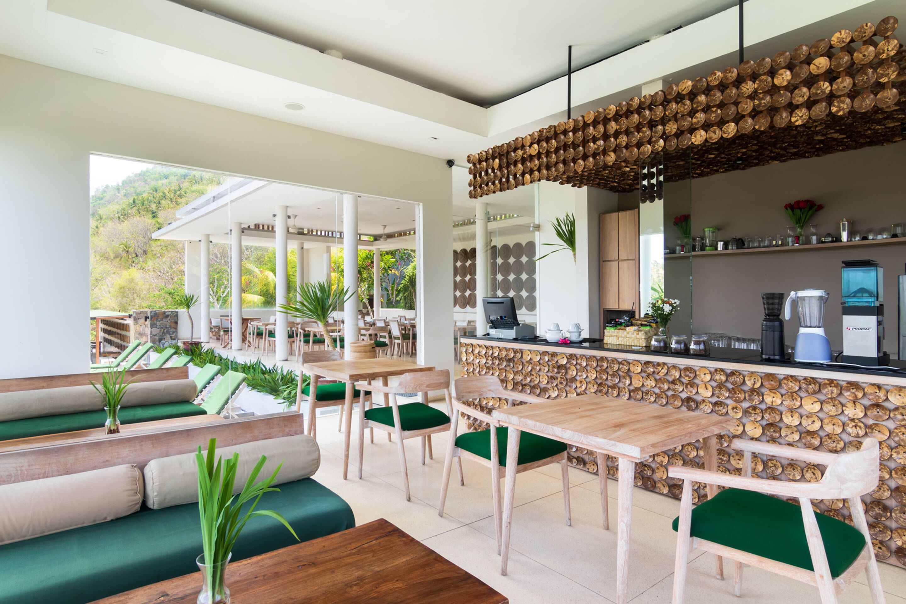 A modern outdoor dining area with wooden tables and green cushioned chairs, a wooden wall with a unique circular pattern, and a view of a lush garden.