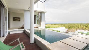 Zona de piscina exterior moderna con vista al océano, terraza de madera y una silla de descanso verde.