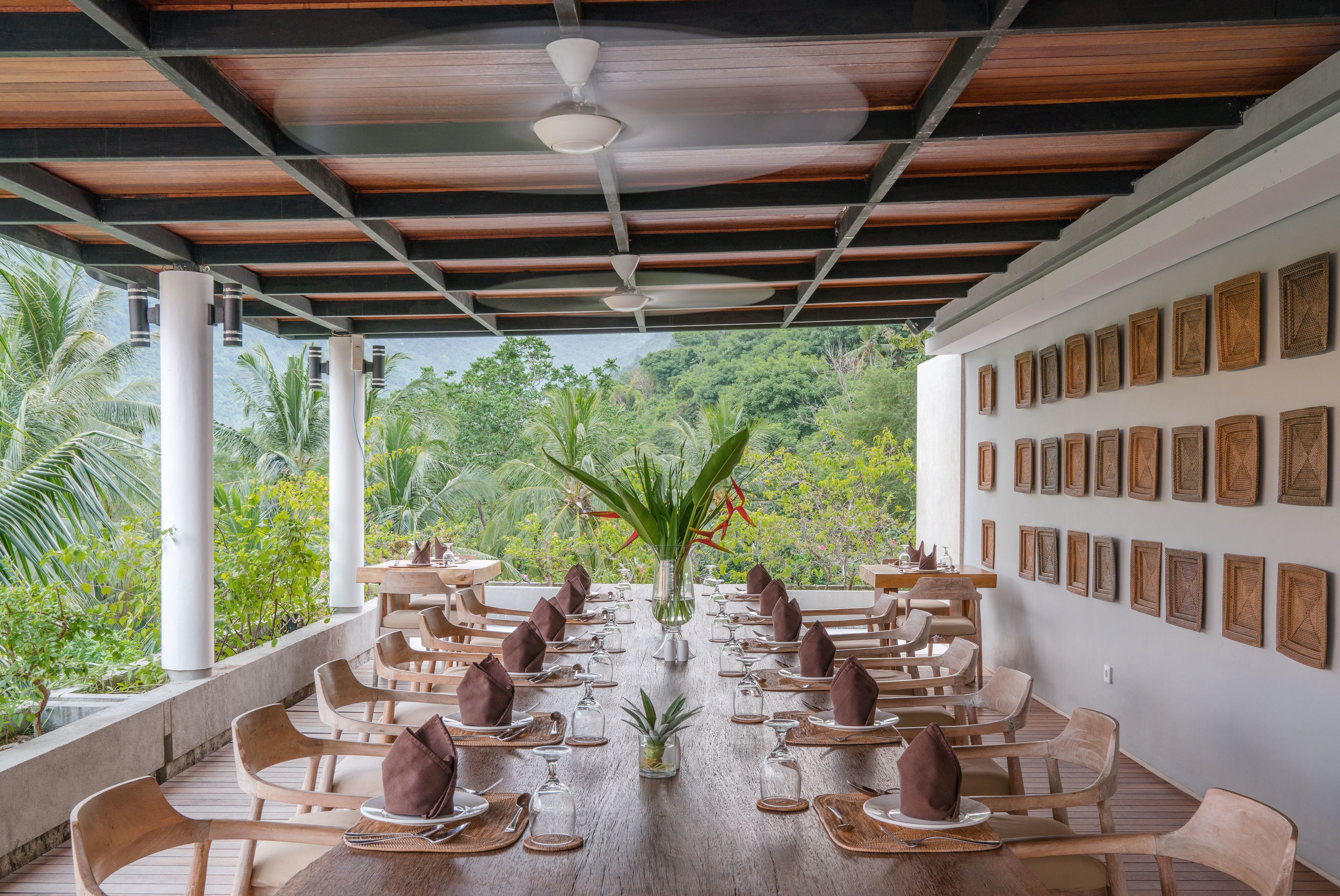 Un área de comedor al aire libre techada, con mesas y sillas de madera, ventilador de techo y obras de arte enmarcadas en las paredes.