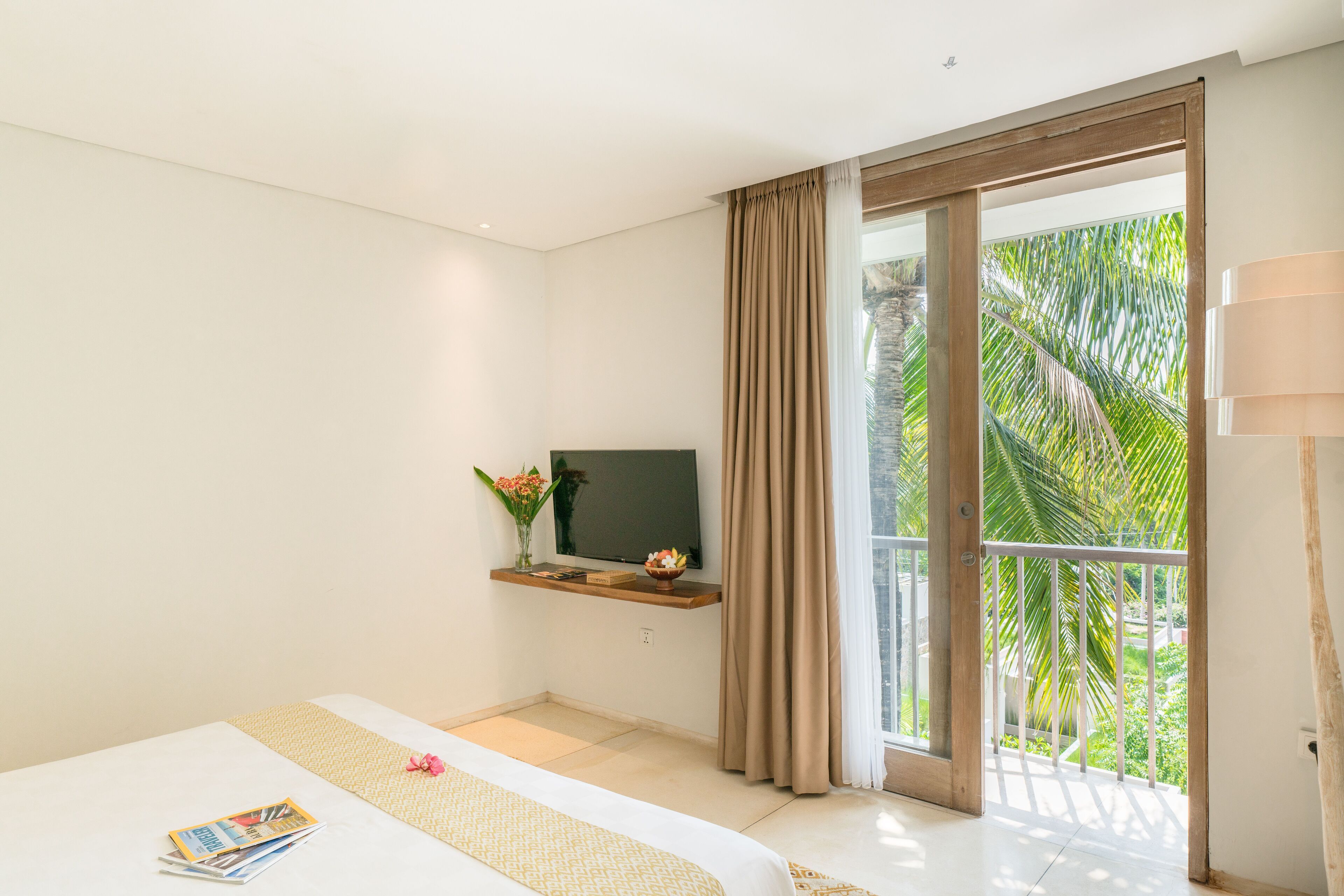 Un dormitorio con cama, televisor en una estantería, balcón con cortinas y vistas a palmeras.