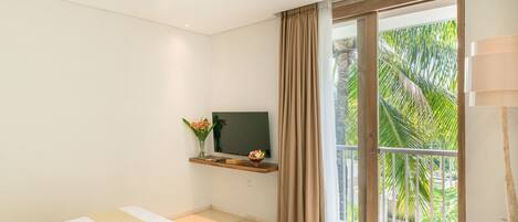Ein Schlafzimmer mit Bett, Fernseher auf einer Ablage, einem Balkon mit Vorhängen und Blick auf Palmen.