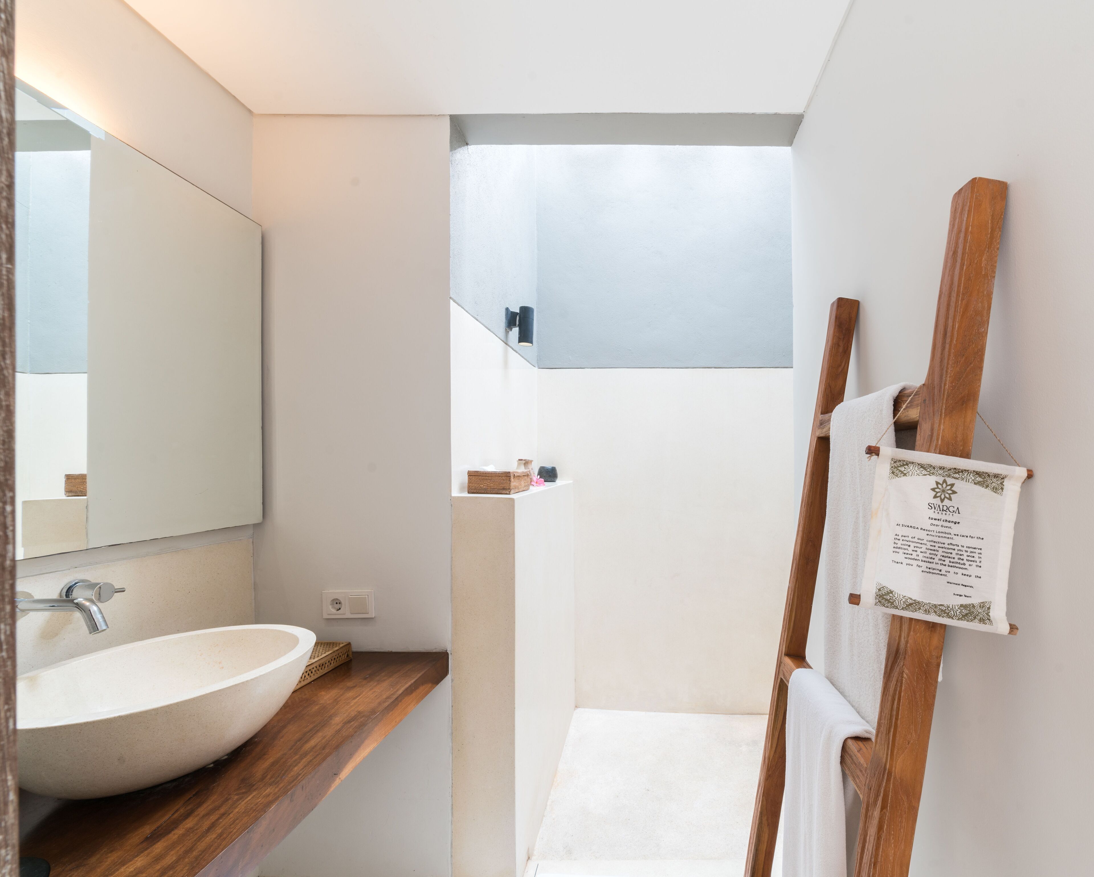 A modern bathroom with a large white sink, wooden countertop, and a mirror. There is a wooden easel with a towel hanging on it and a small shelf with toiletries.