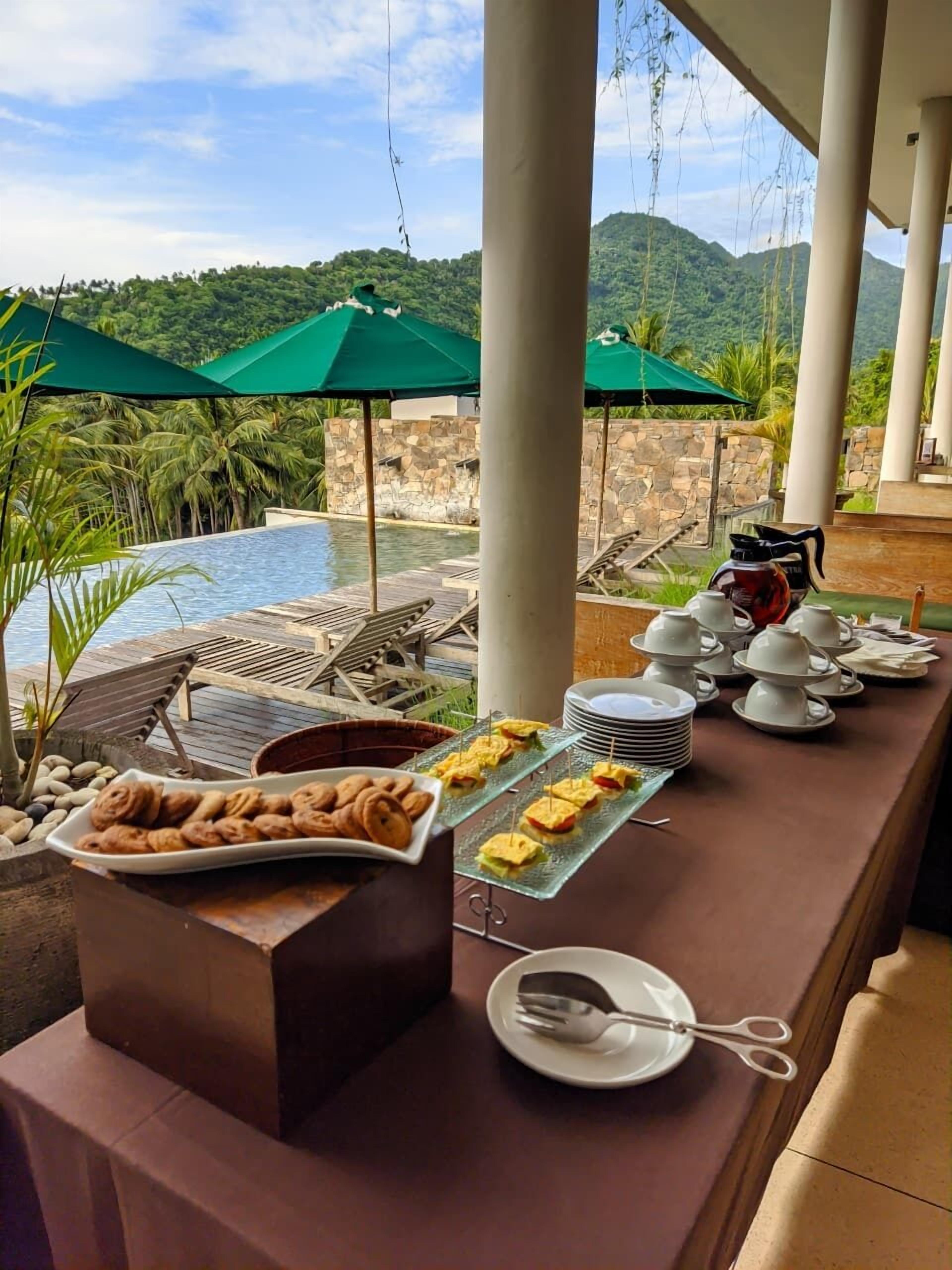 Un bufé de desayuno con pastelería y una estación de café, con vistas a una piscina y exuberante follaje.