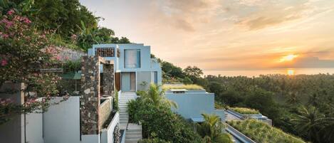 A modern house with a staircase leading to a balcony, surrounded by lush greenery and a view of the sunset over the ocean.