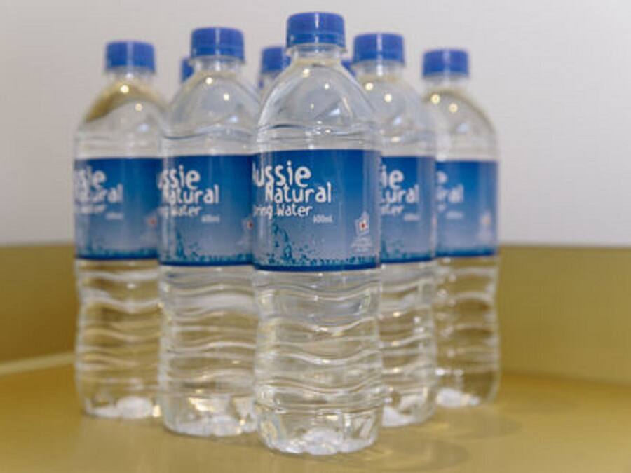 Une rangée de bouteilles en plastique transparent, dotées de bouchons bleus, sur lesquelles on peut lire l’étiquette « Wessie, eau de source naturelle », le tout placé sur une surface réfléchissante.