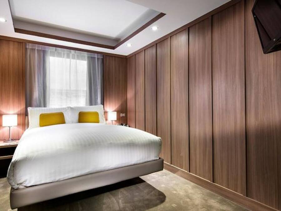 Een moderne hotelkamer met een groot bed, houten lambrisering, een nachtkastje met een lamp en een groot raam met gordijnen.