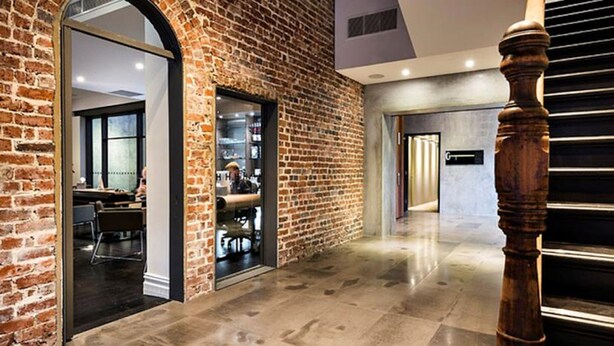 Un pasillo con pared de ladrillo, un pilar de madera y una puerta de cristal que da a una habitación con escritorio y sillas.