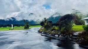 Un camino pavimentado que lleva a una casa rodeada de exuberante vegetación y una montaña al fondo.