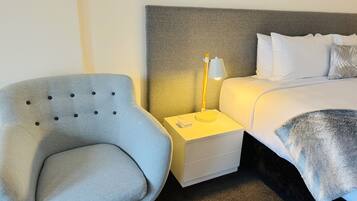 A neatly arranged hotel room with a grey upholstered armchair, a white bedside table with a lamp, and a neatly made bed with white linens.