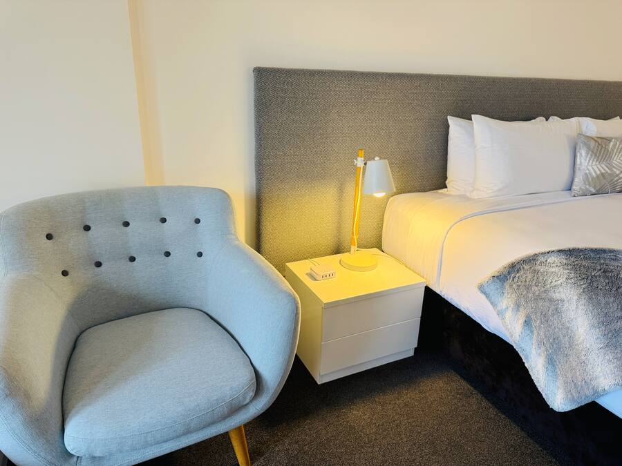 A neatly arranged hotel room with a grey upholstered armchair, a white bedside table with a lamp, and a neatly made bed with white linens.