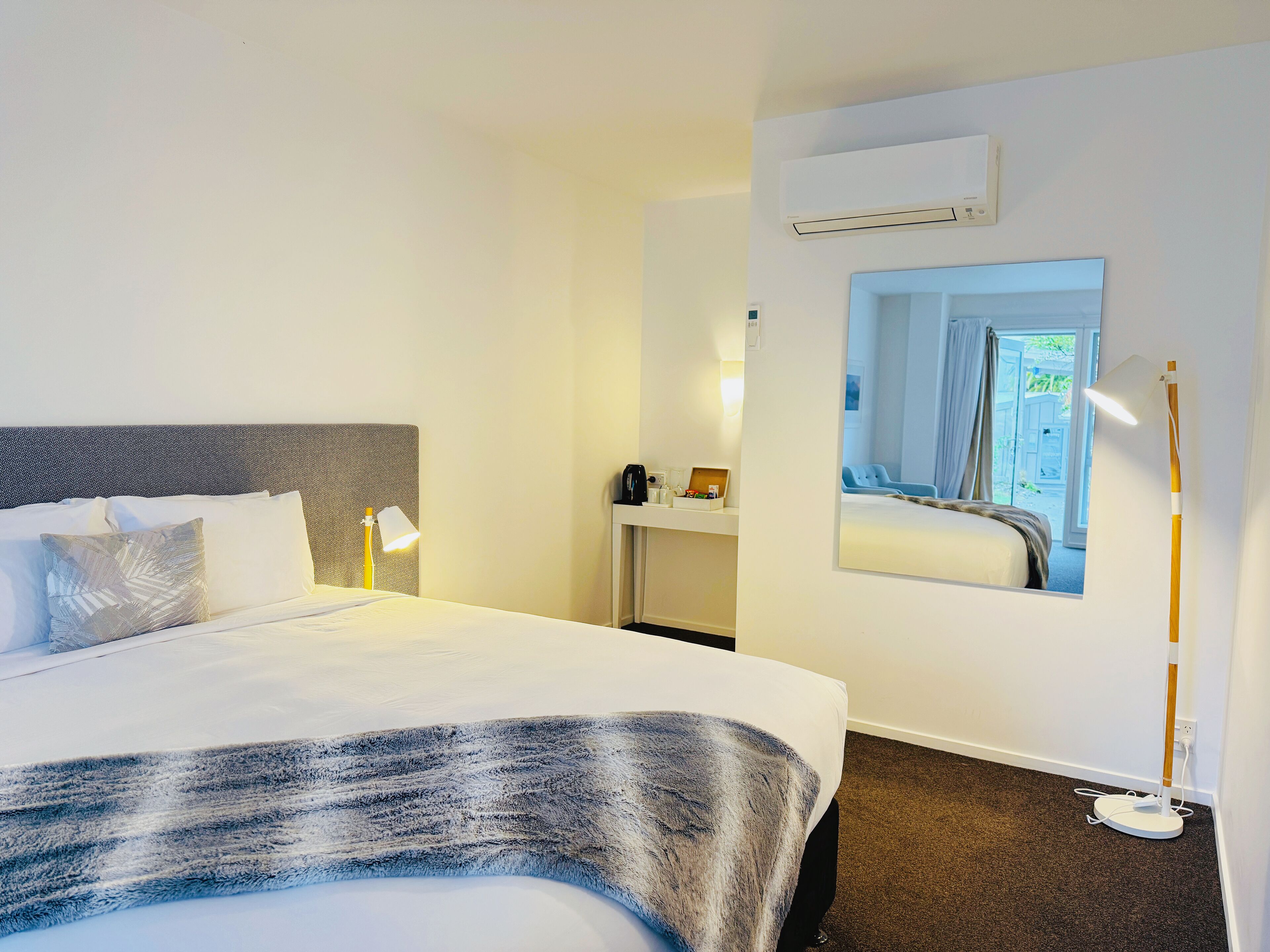 Ein Hotelzimmer mit Bett, Nachttisch, Lampe, Klimaanlage und einem Spiegel, der den Raum wiederspiegelt.