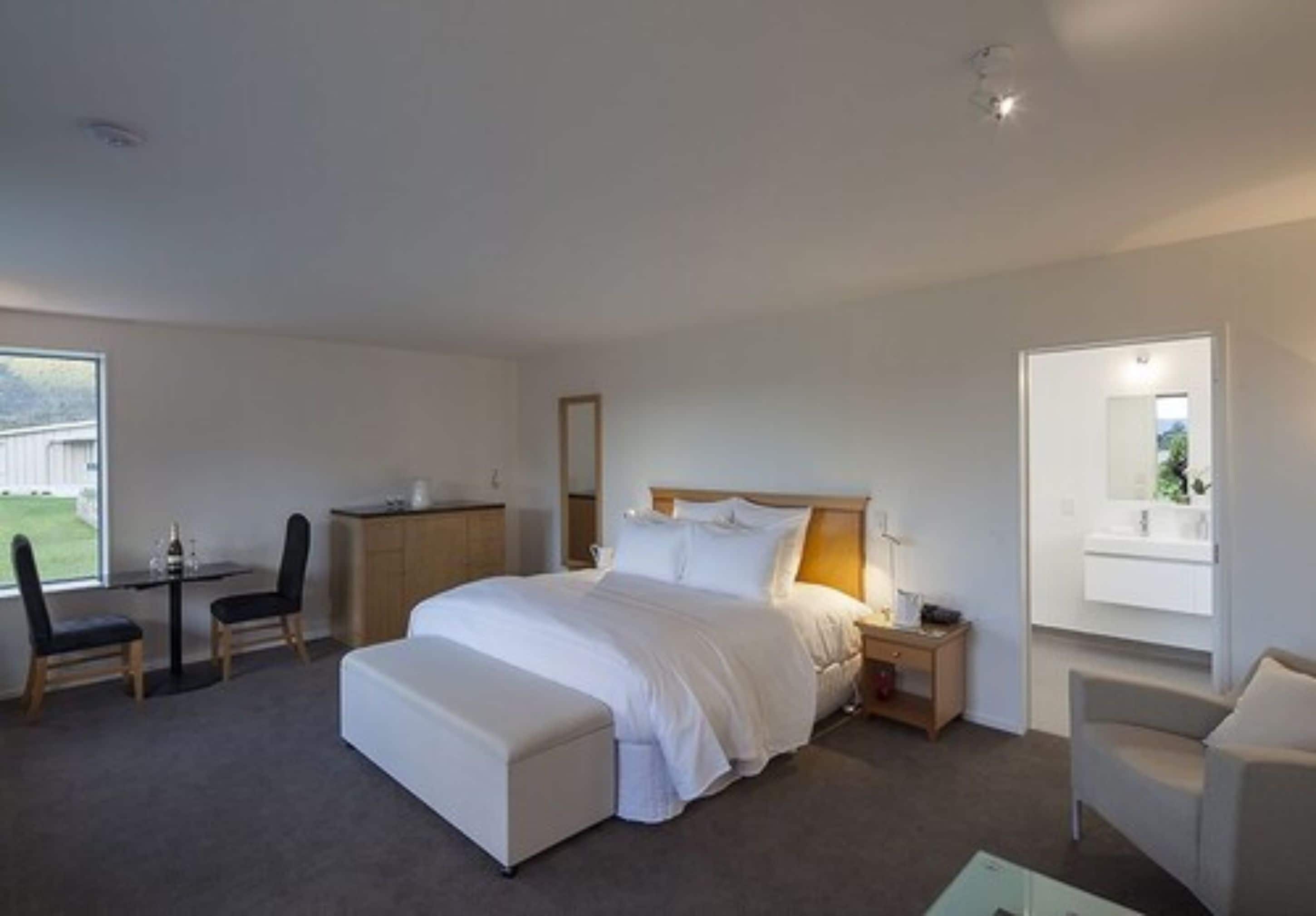 A modern hotel room with a large bed, a desk, and a chair. There is a small table with a lamp, a mirror, and a window with a view of the outdoors.