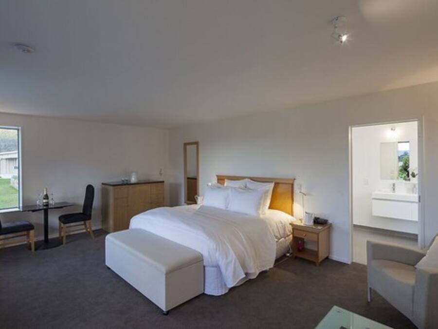 A modern hotel room with a large bed, a desk, and a chair. There is a small table with a lamp, a mirror, and a window with a view of the outdoors.