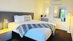Una habitación de hotel con una cama grande, una mesita de noche con lámpara, un tocador y un equipo de aire acondicionado.