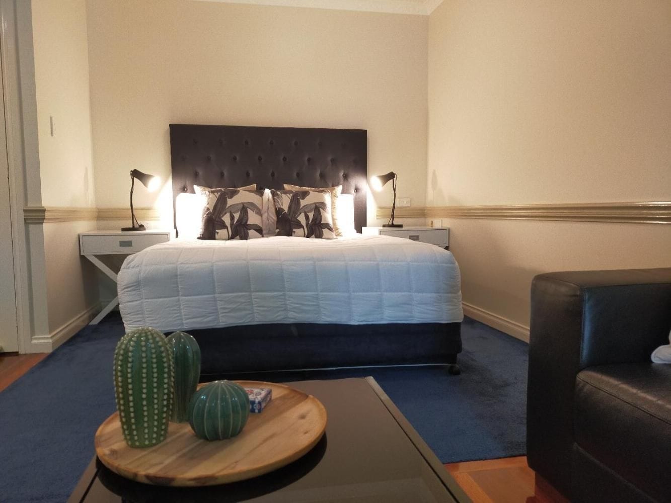Una habitación de hotel con una cama grande, dos lámparas de noche, una mesita de madera con adornos y un sillón de cuero oscuro.