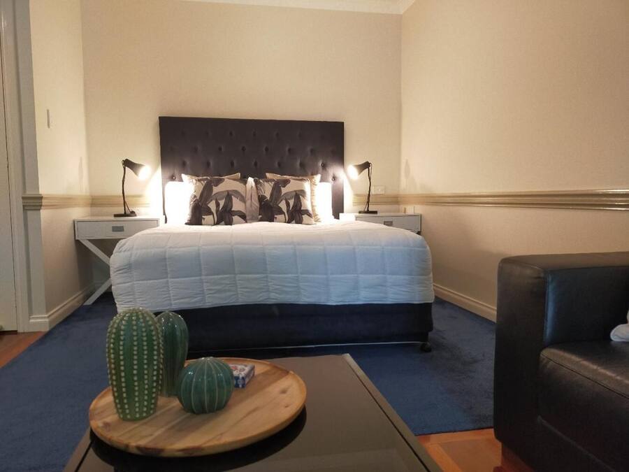 Una habitación de hotel con una cama grande, dos lámparas de noche, una mesita de madera con adornos y un sillón de cuero oscuro.