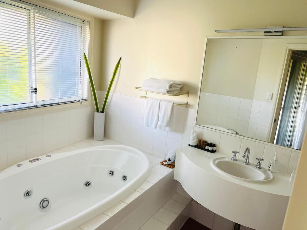 A modern bathroom with a bathtub, two sinks, a large mirror, and a window with blinds.