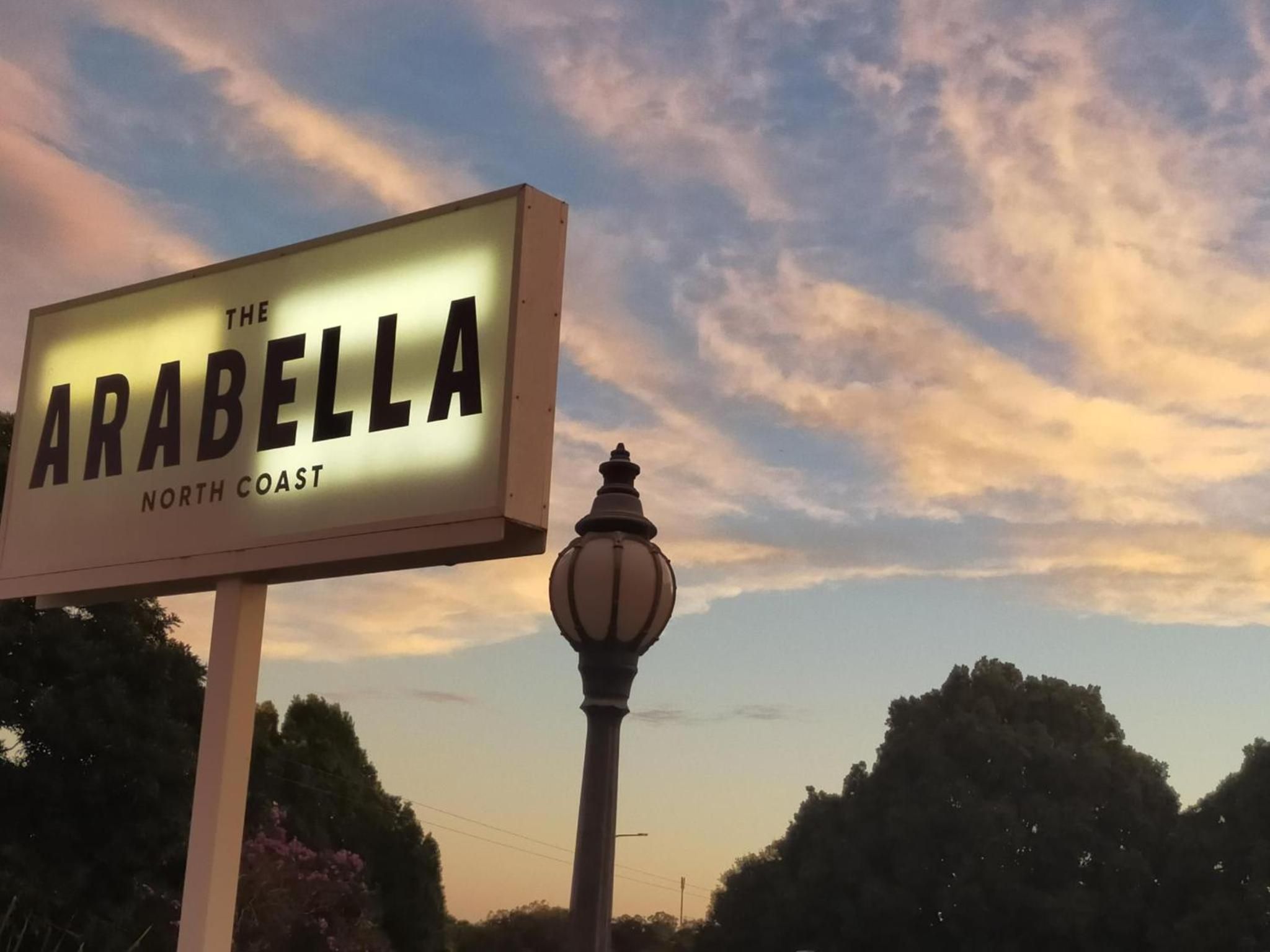 Schild für 'The Arabella North Coast' mit einer Straßenlaterne und Bäumen im Hintergrund.