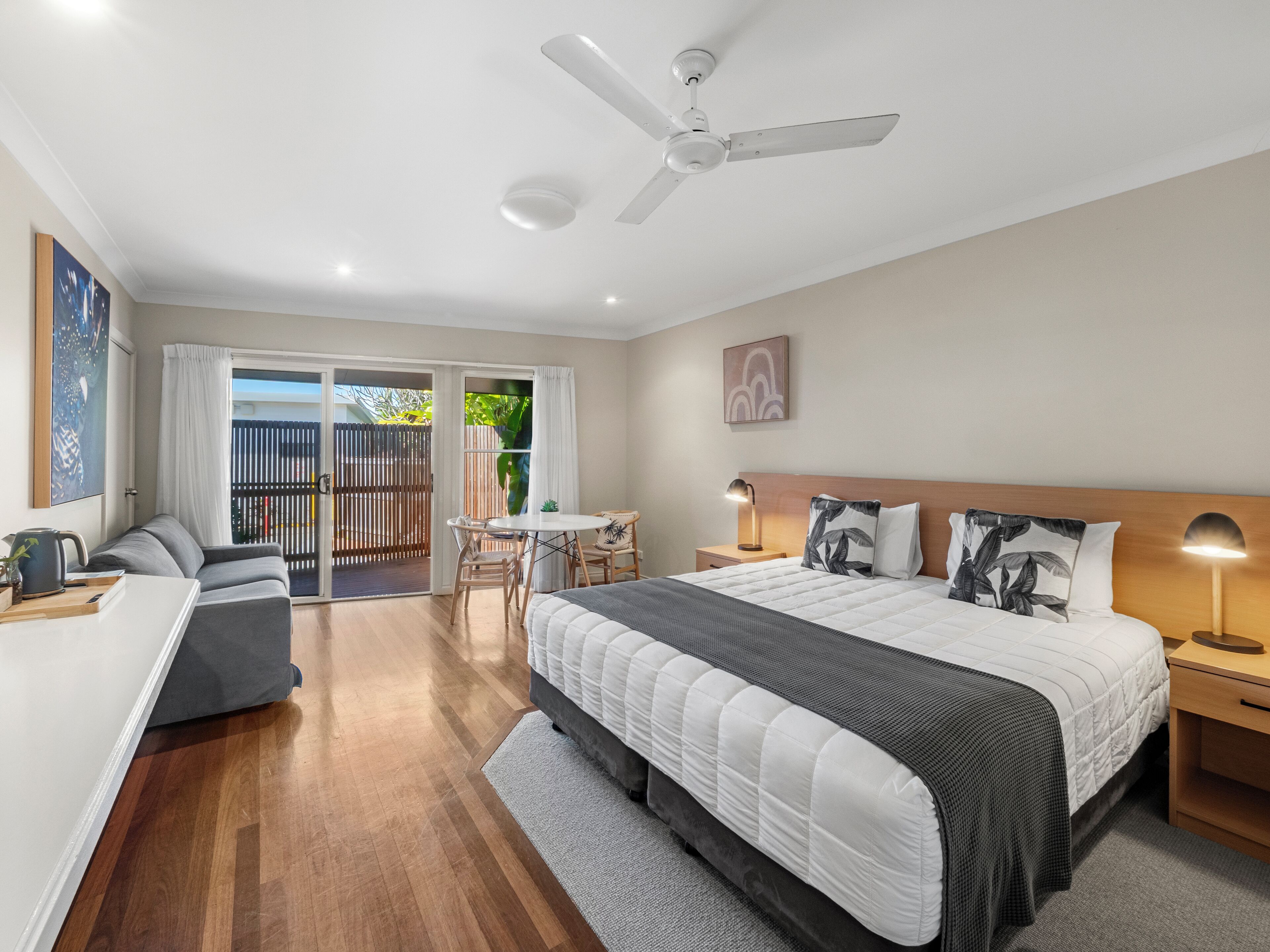 Une chambre d’hôtel moderne équipée d’un lit, de tables de chevet, d’un bureau, d’un ventilateur de plafond et d’une porte-fenêtre coulissante donnant sur un espace extérieur.