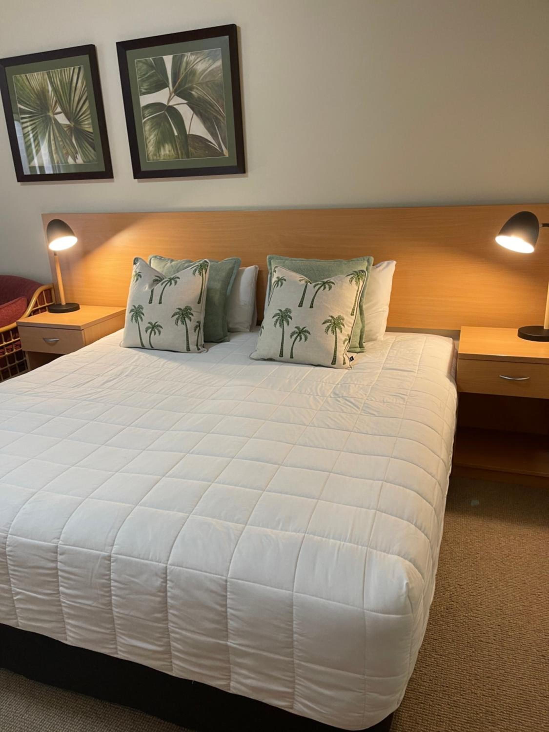 A neatly made bed with a quilted white duvet, two bedside lamps, and two framed tropical leaf artworks on the wall.