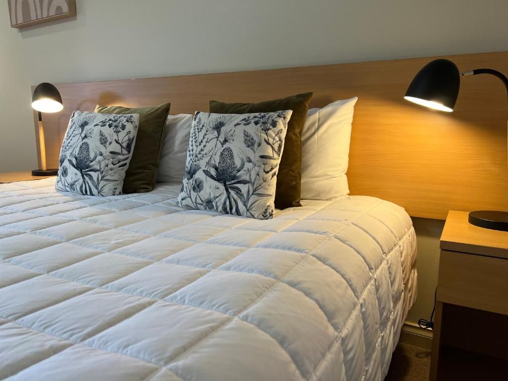 A neatly made bed with a white duvet and floral patterned pillows, a wooden headboard, and two bedside lamps on a nightstand.
