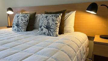 A neatly made bed with a white duvet and floral patterned pillows, a wooden headboard, and two bedside lamps on a nightstand.