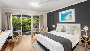 A bedroom with a bed, a desk, and a chair. There is a window with curtains and a view of greenery. - Arabella Garden Inn Motel (Wollongbar)