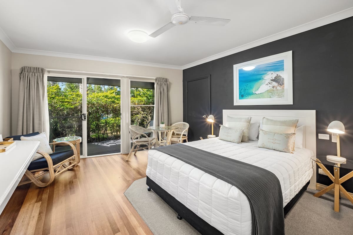 A bedroom with a bed, a desk, and a chair. There is a window with curtains and a view of greenery.