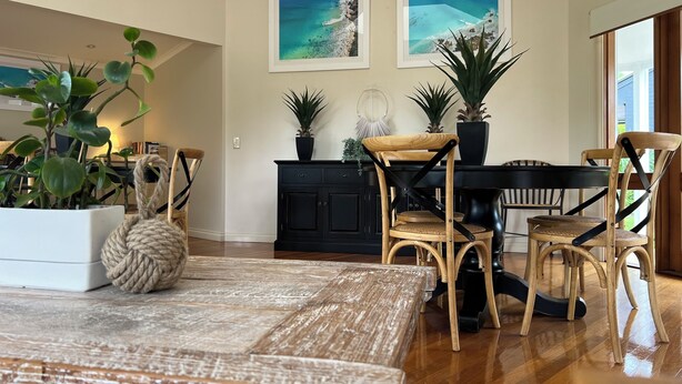 A dining area with a wooden table, chairs, and a black cabinet. There are two framed pictures on the wall, and a potted plant on the cabinet. - Arabella Garden Inn Motel (Wollongbar)