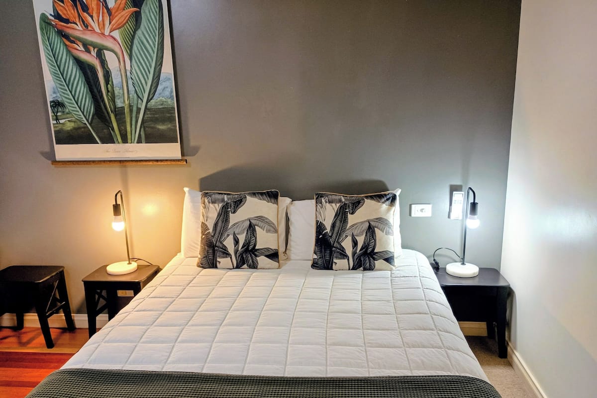 A neatly made bed with a white quilt, two pillows with a leaf pattern, a small bedside table with a lamp, and a wall-mounted light fixture.