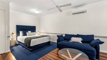 A bedroom with a bed, a blue sofa, a glass coffee table, a ceiling fan, and an air conditioning unit.