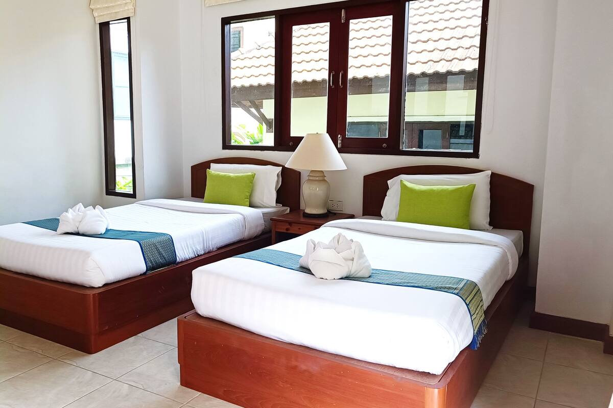 Habitación de hotel con dos camas, cada una con ropa de cama blanca y almohadas verdes, cabecero de madera, mesita de noche con lámpara y ventana con vista a edificios.