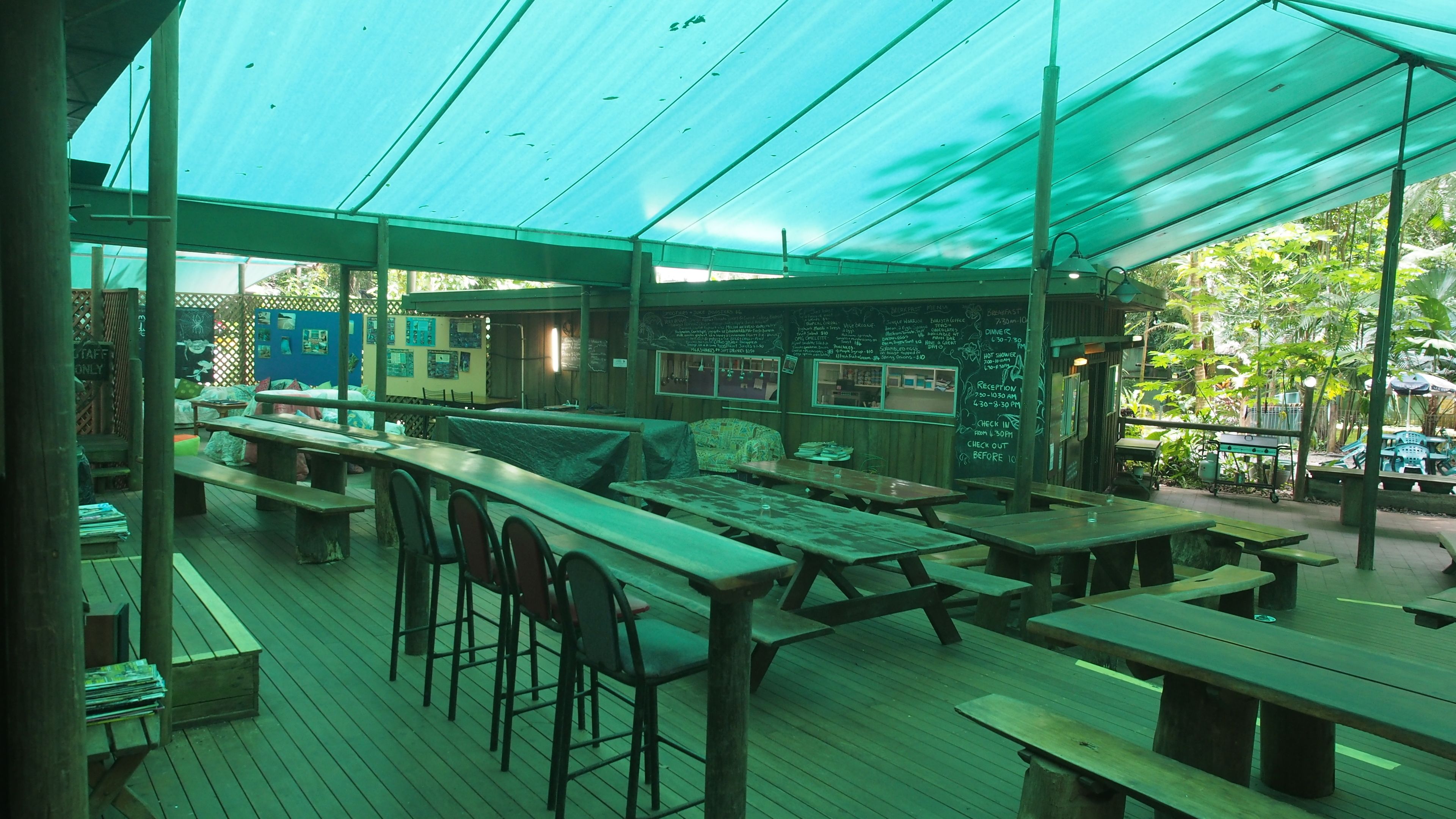 An outdoor restaurant with wooden tables and chairs, covered by a green canopy.