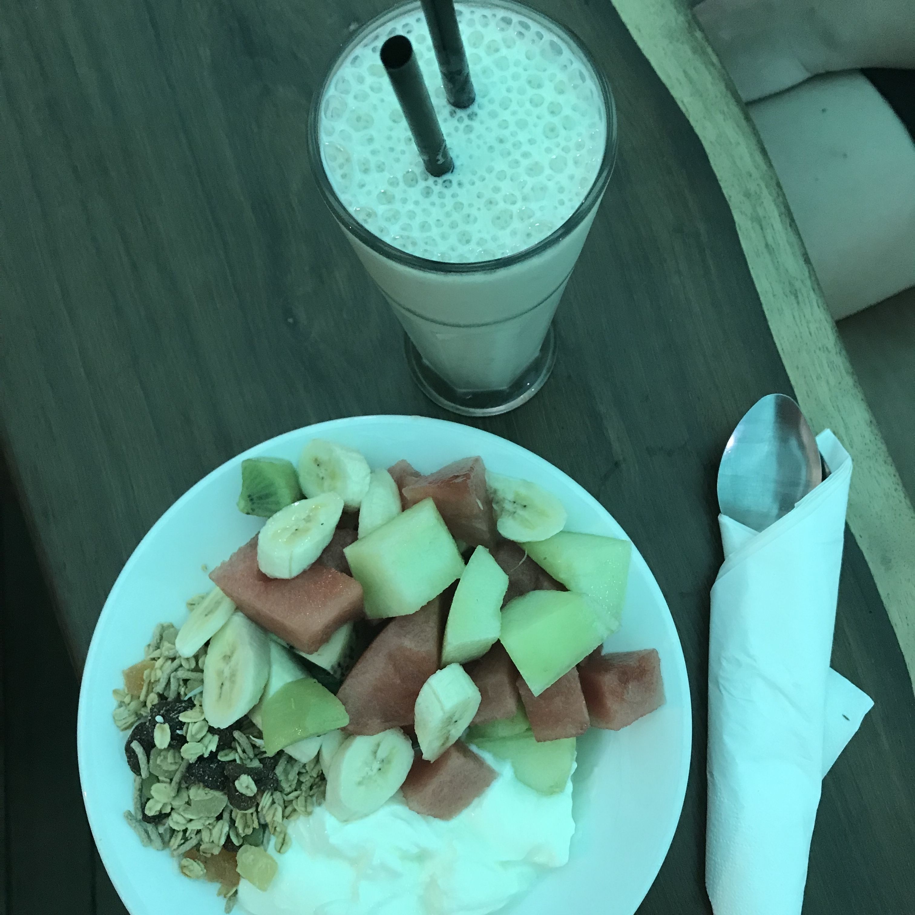 Un plato de fruta y granola, un vaso de batido y una cuchara sobre una mesa.