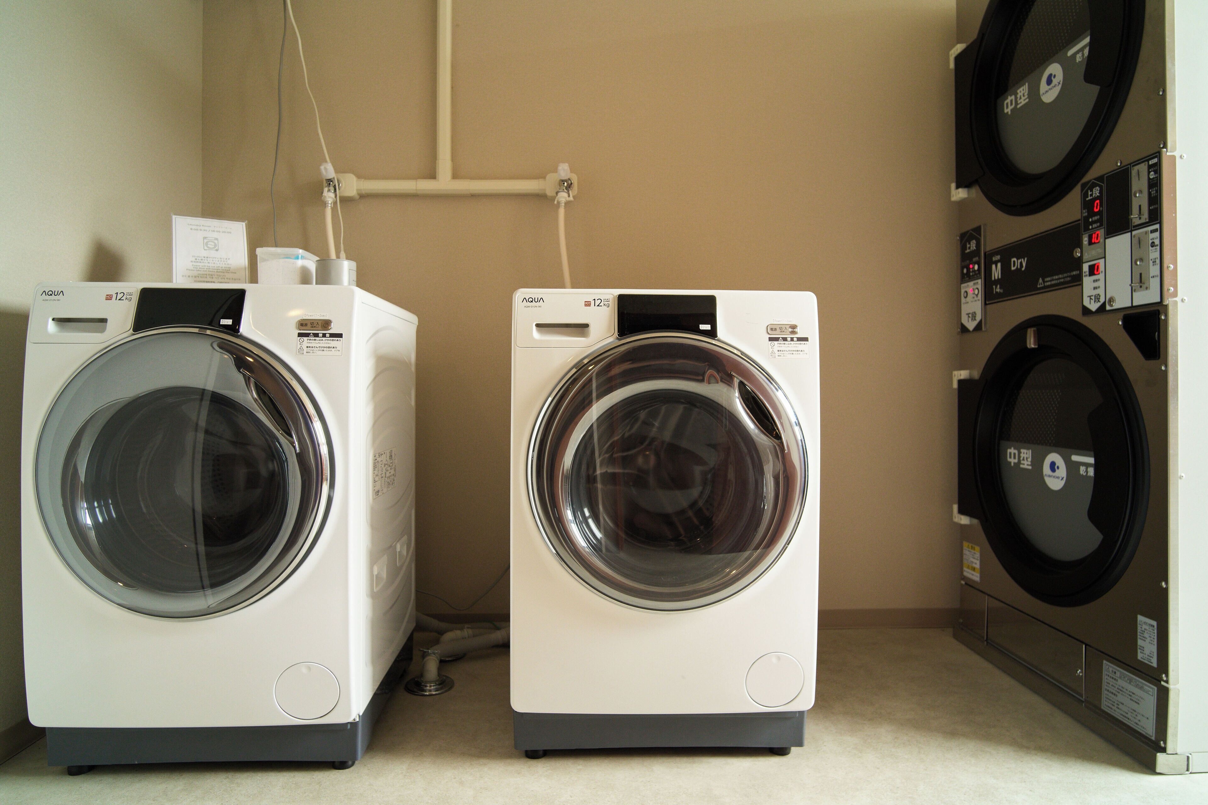 Two front-loading washing machines in a laundry room with a control panel and a wall-mounted dryer.