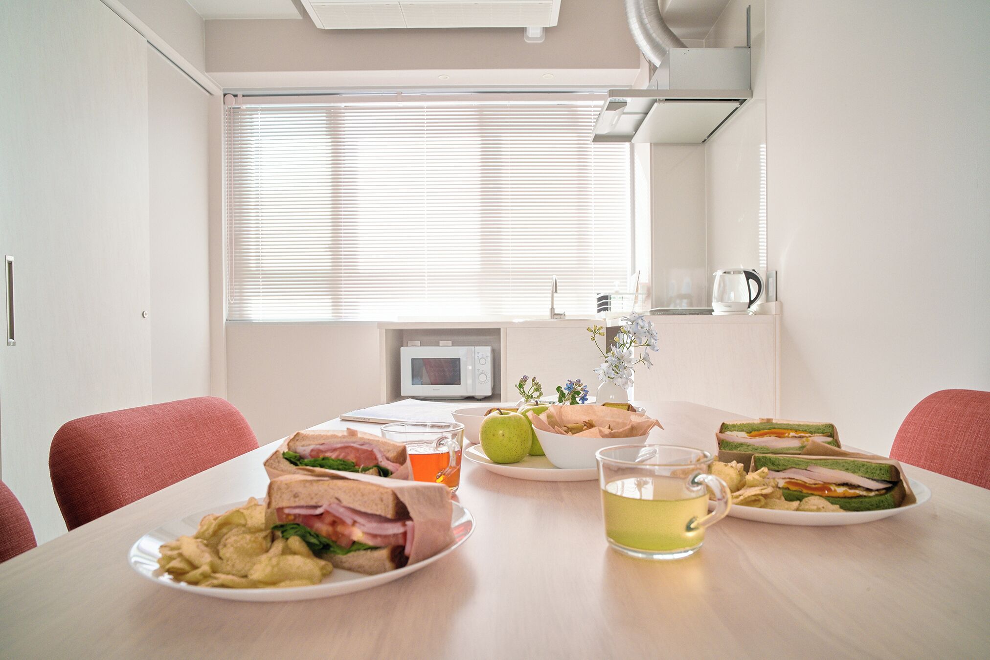 A dining table with sandwiches, apples, and a cup of tea in a bright room with a window and a microwave in the background.